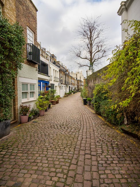 A narrow cobblestone street in a residential area with tightly packed terraced houses on both sides. Some houses feature small balconies with black metal railings and windows with white frames. Potted