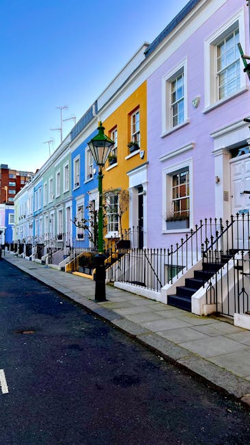 A narrow cobblestone street in a residential area with tightly packed terraced houses on both sides. Some houses feature small balconies with black metal railings and windows with white frames. Potted